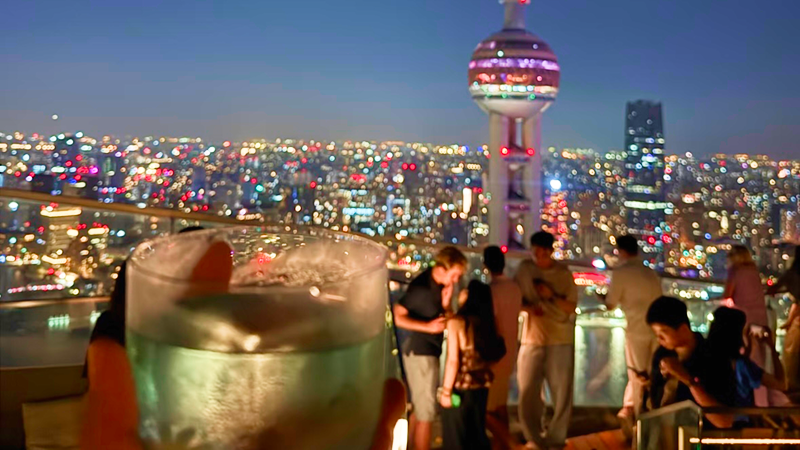 Shanghai skyline at night viewed from a rooftop bar, with a cocktail in the foreground and the Oriental Pearl Tower glowing over the city lights.