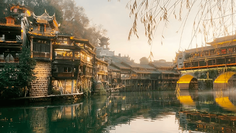 At dusk in Fenghuang Ancient Town, as the sun dips below the horizon and the lights begin to flicker on, the river mirrors the riverside buildings, a scene both cozy and enchanting.