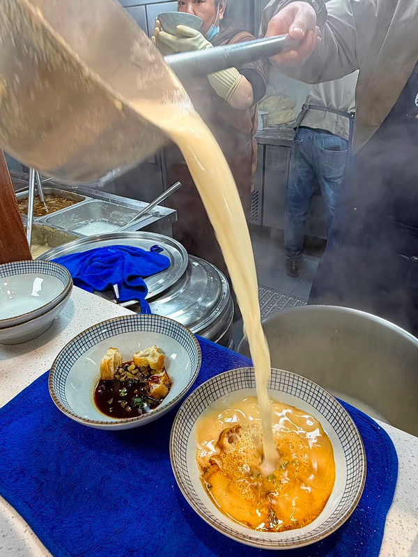 Fresh soy milk being poured into a bowl of savory douhua at a Hangzhou breakfast stall
