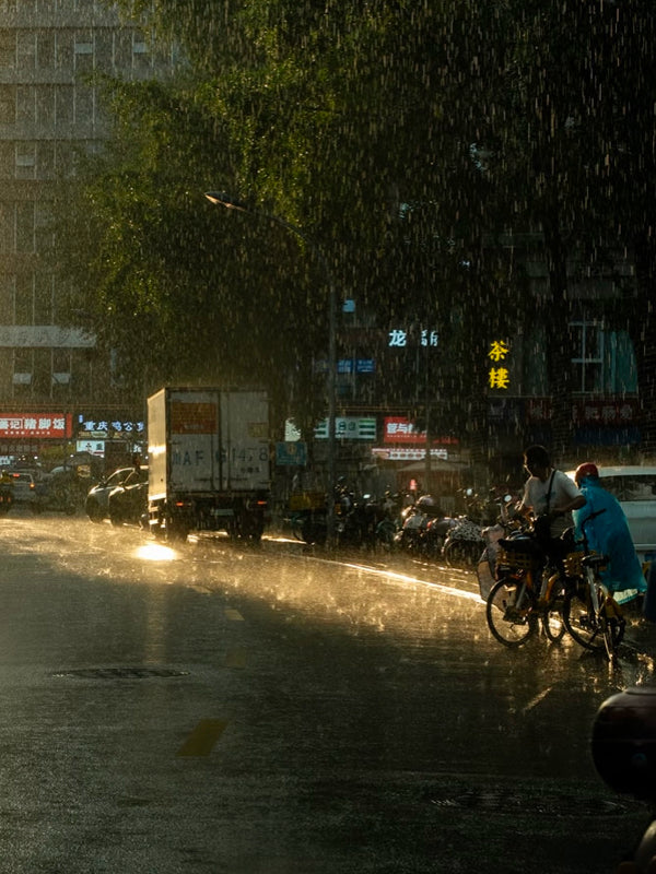 Local cyclists riding through a rainy street in Chengdu under golden streetlights, reflecting daily life in all weather conditions
