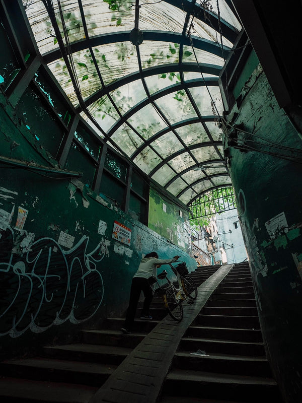 Local resident pushing a shared bike up a dimly lit urban underpass in a gritty Wuhan alley, capturing the raw neighborhood energy