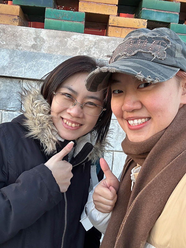 Two women dressed in winter clothing giving thumbs-up during a local walk in a Beijing neighborhood, showcasing hospitality and connection