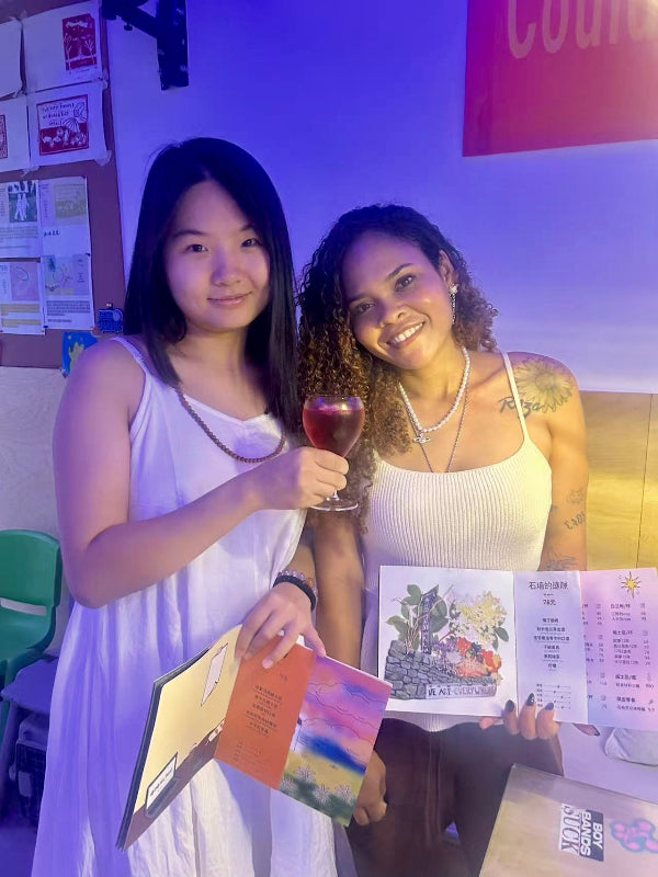 Two women enjoying Chengdu nightlife at a zine-friendly local bar, celebrating creative expression with handmade booklets and drinks