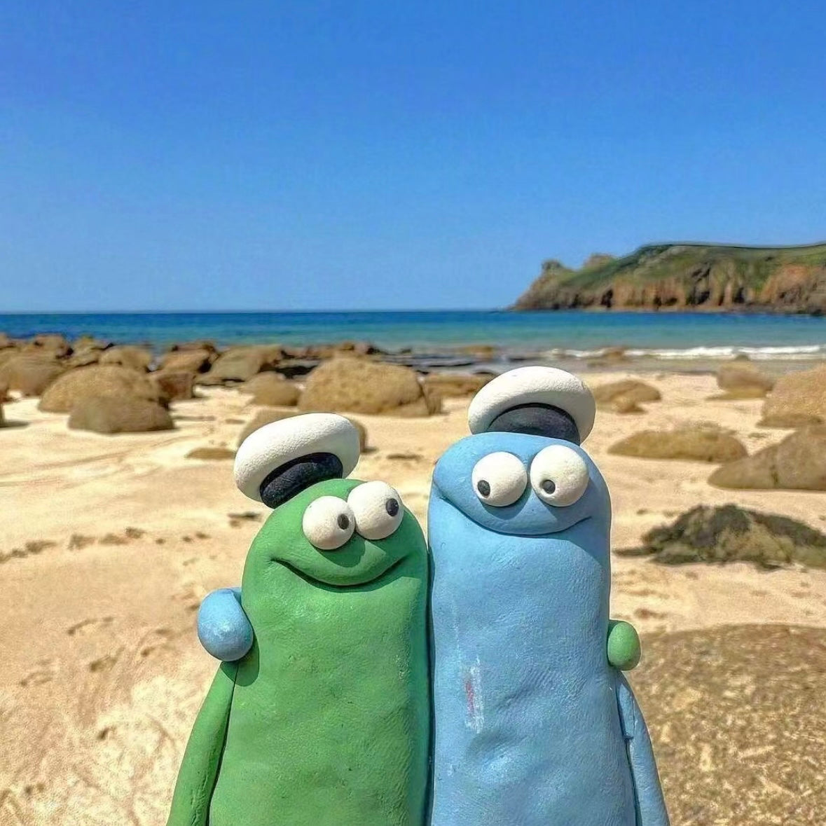 Two clay cartoon figures hugging on a sandy beach with a blue sky, symbolizing friendship, gratitude, and global connections.