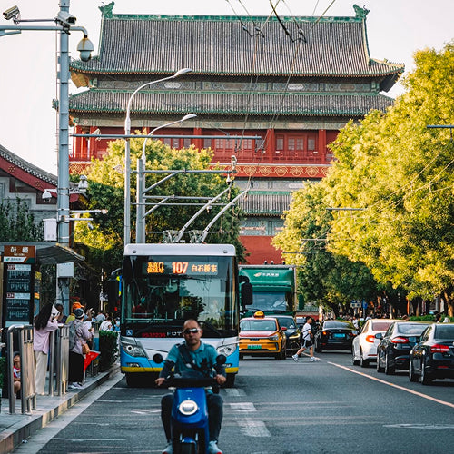 Bustling Beijing street scene with local bus and scooter riders near historic Drum Tower in a vibrant neighborhood setting