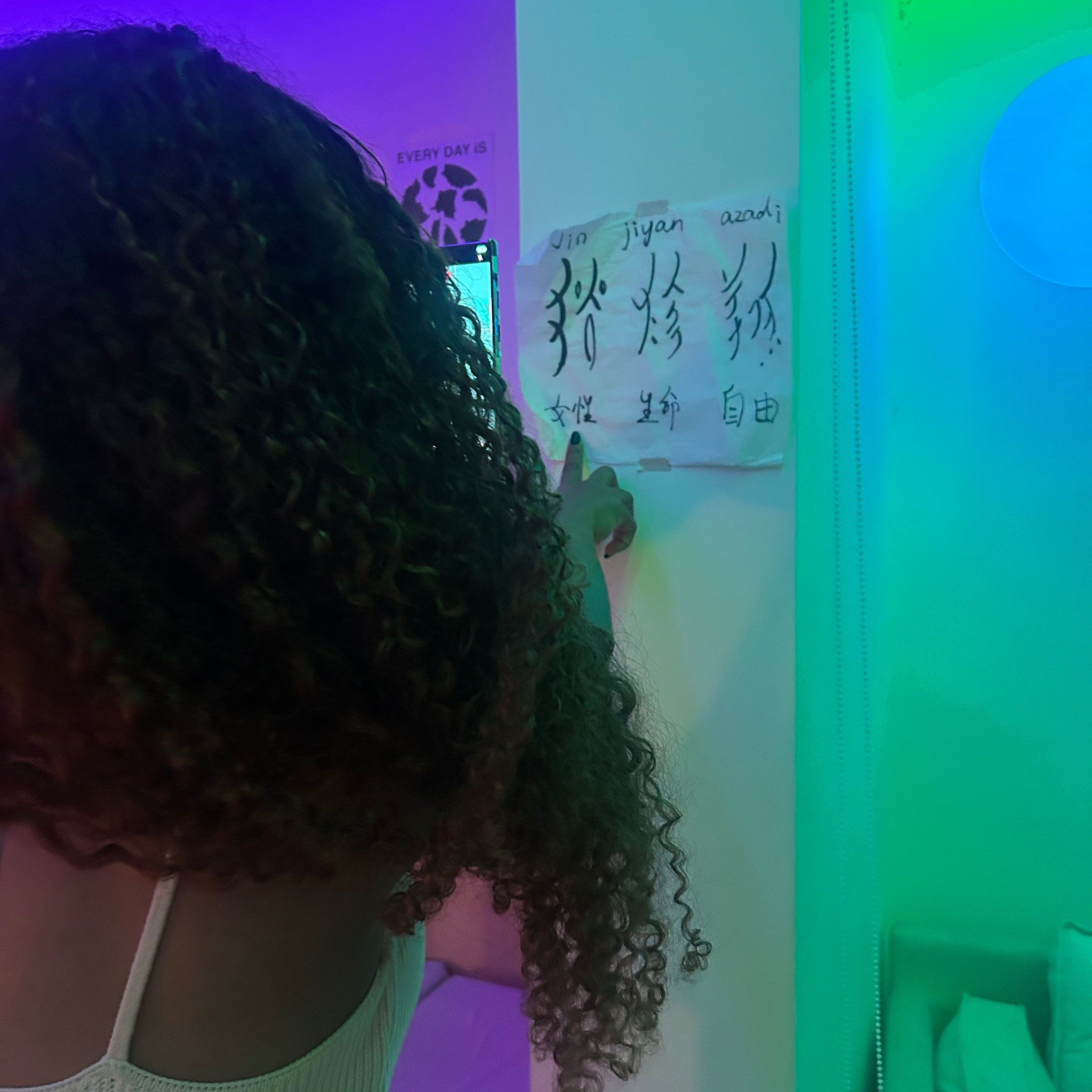 Young woman pointing at calligraphy on a neon-lit wall featuring the Nvshu calligraphy 'Female, Life, Freedom' with Chinese translation.